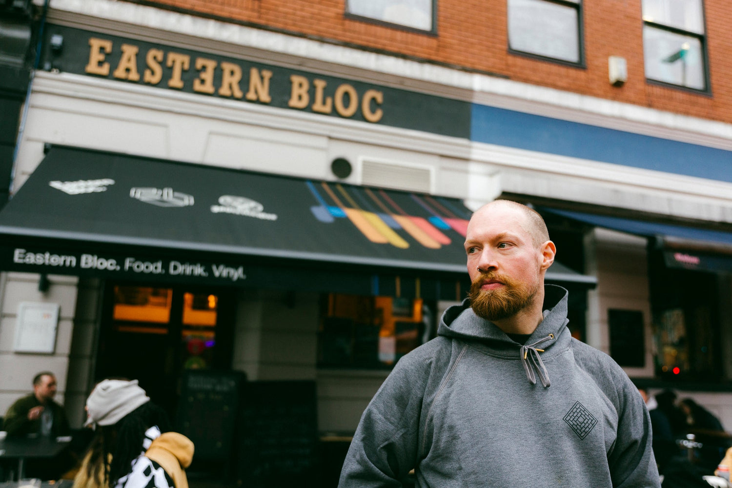 Bearded man wearing gray hoodie standing outside Eastern Bloc cafe with black awning and brick facade