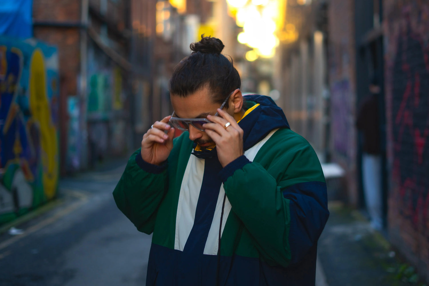 Man in green jacket adjusting sunglasses in urban alley with street art and graffiti walls.