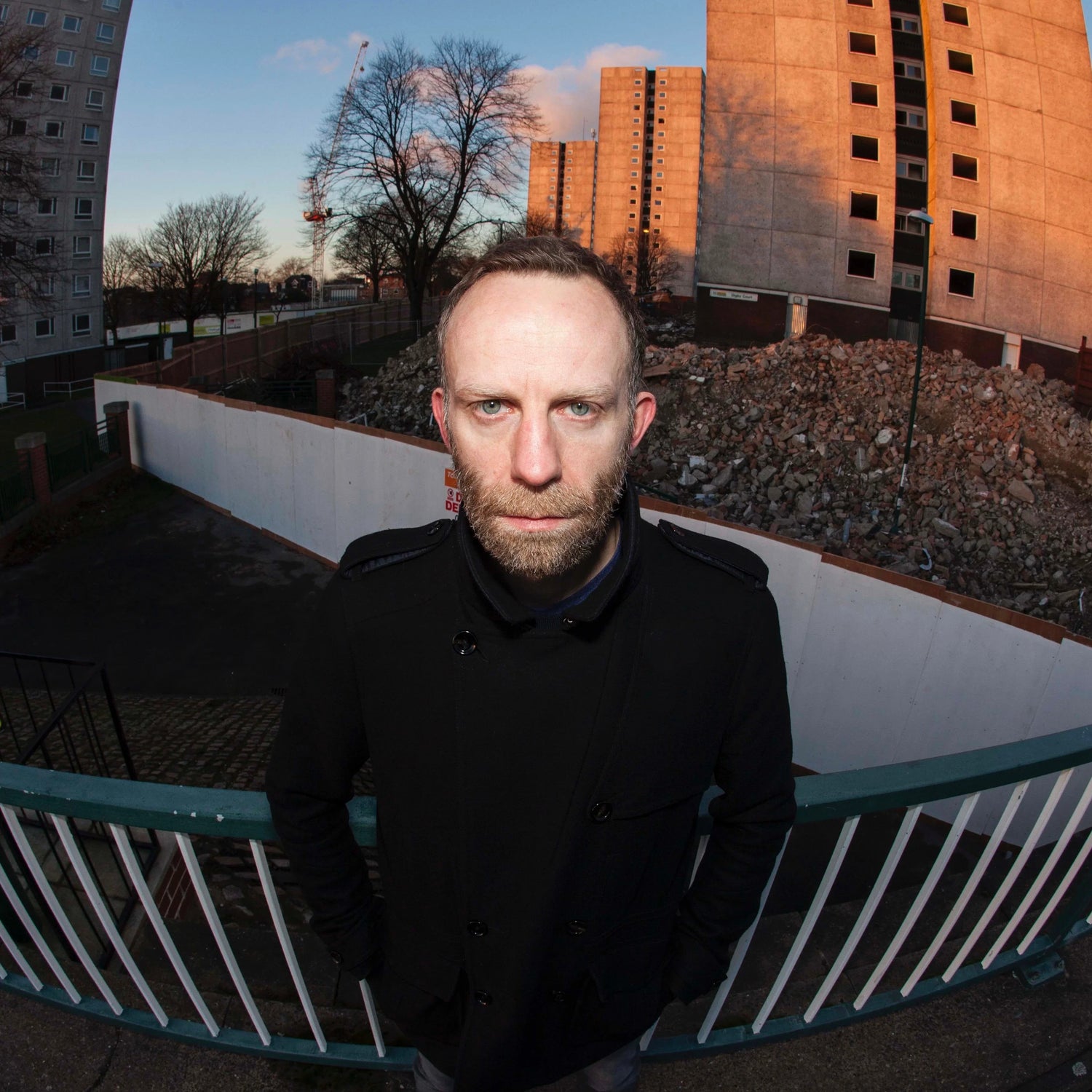 Man in black coat standing in front of urban ruins and high-rise buildings at sunset