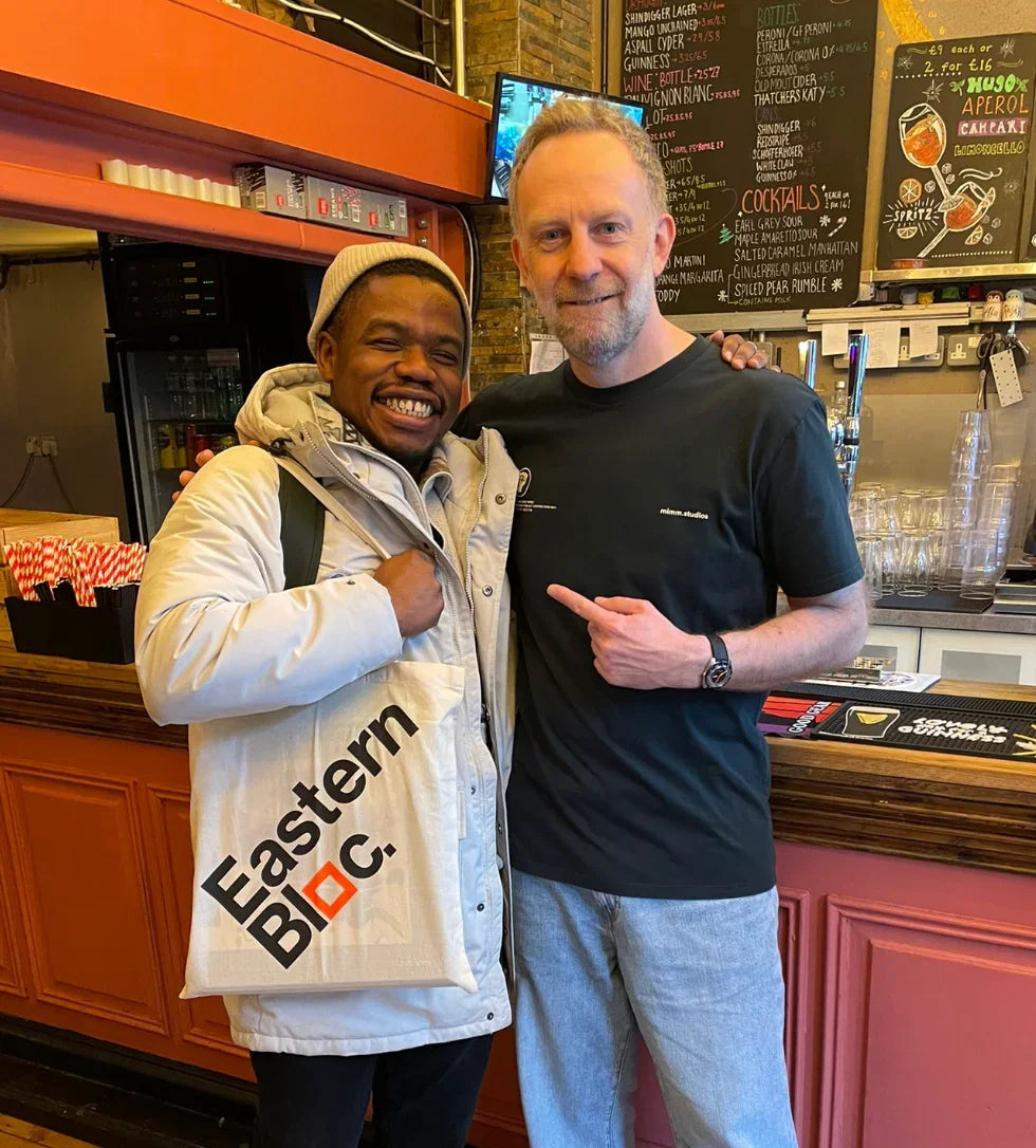 Two men smiling indoors, one holding an Eastern Bloc Records tote bag by a bar counter