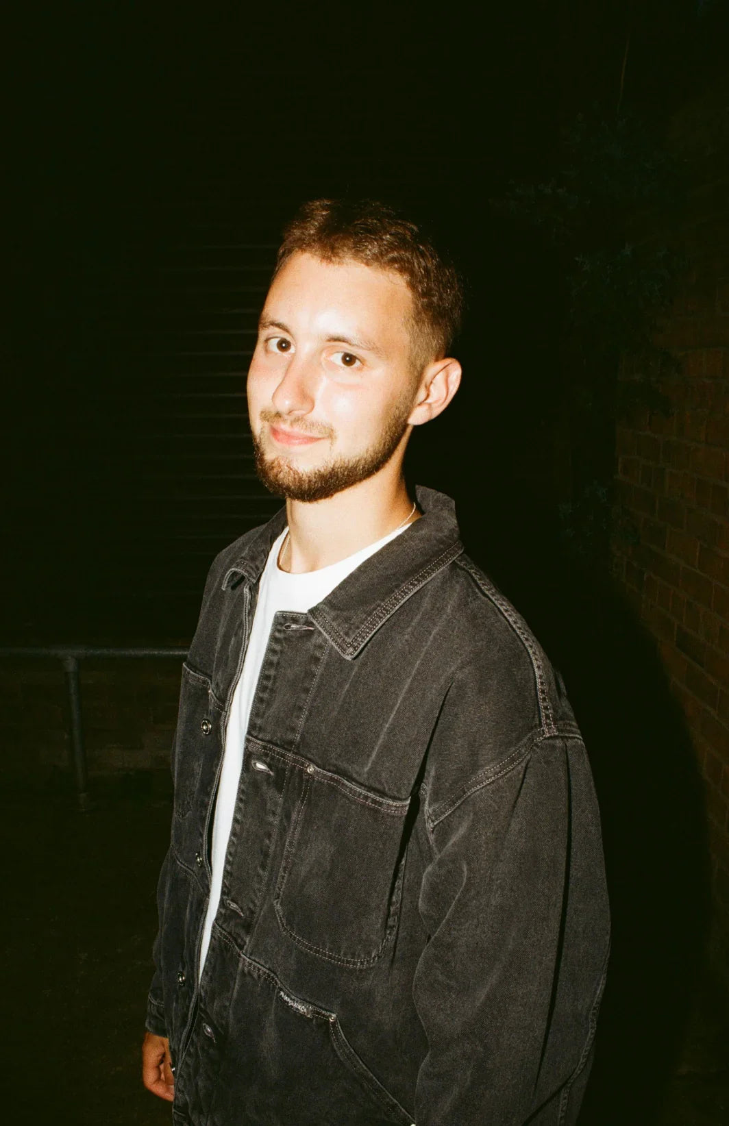 Young man in a black denim jacket standing outdoors at night with brick wall background