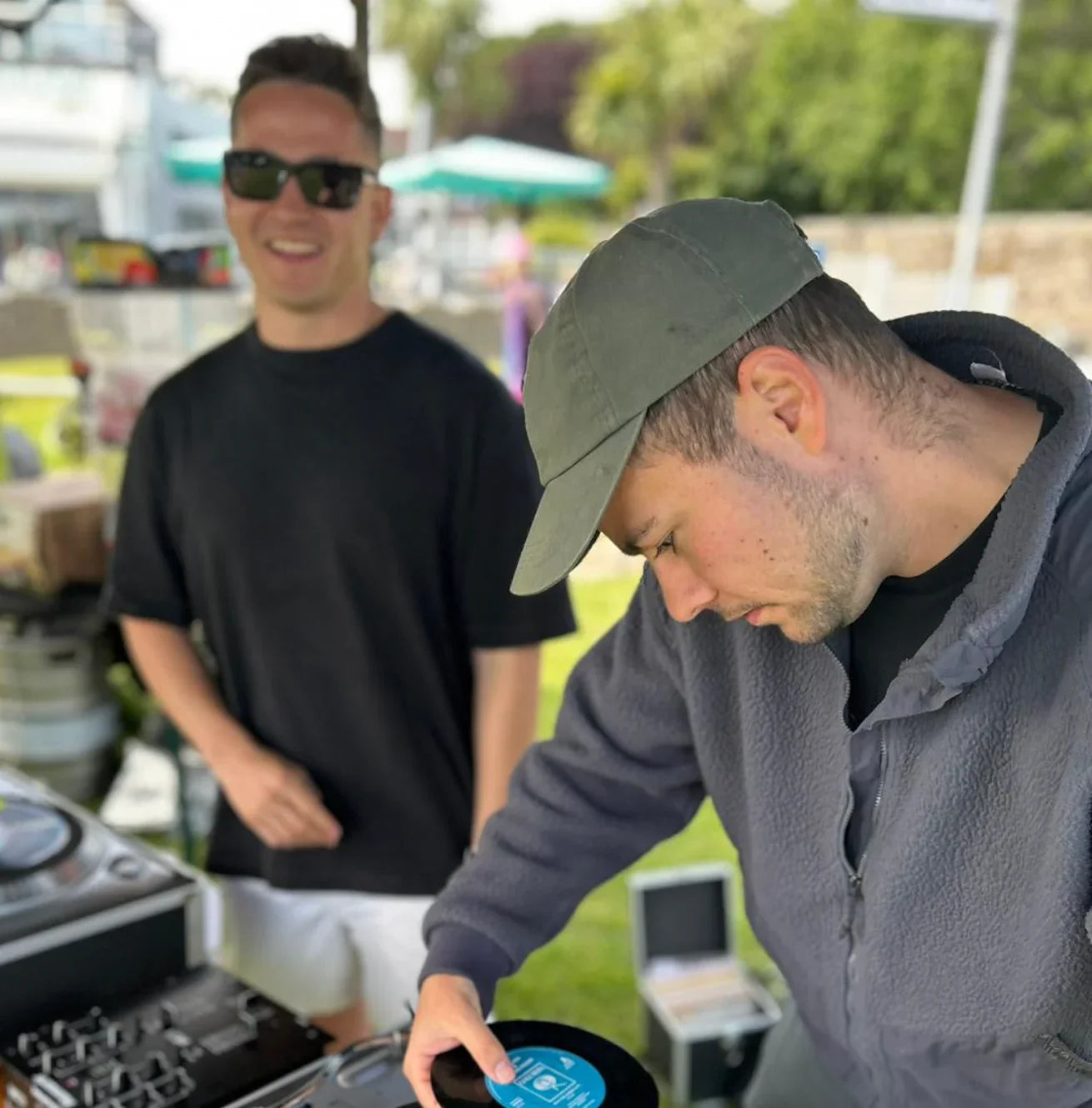 Two men at an outdoor vinyl DJ setup, one placing a record on a turntable.