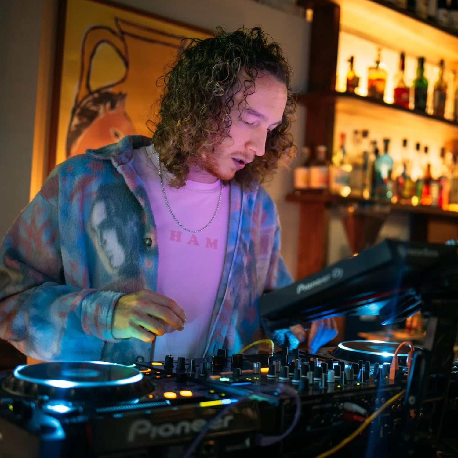 DJ with curly hair mixing music on Pioneer turntables in a bar with colorful lighting