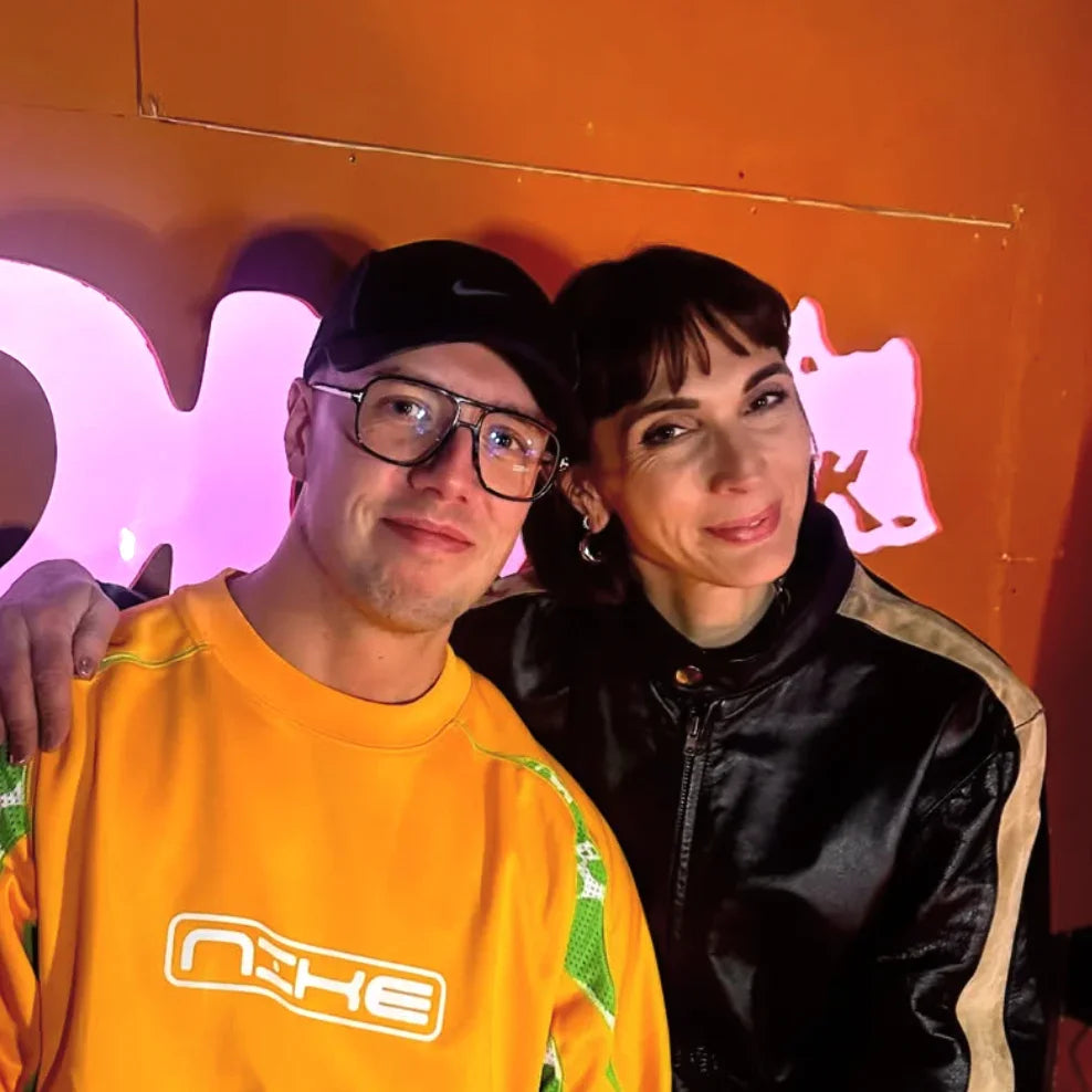 Two people posing together in front of an orange wall with neon lighting, wearing casual streetwear.