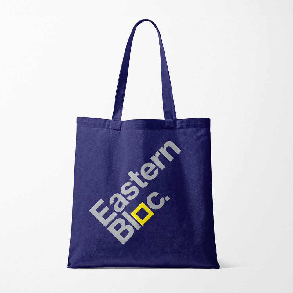 Navy blue tote bag with diagonal white text 'Eastern Bloc.' and a yellow square detail on white background