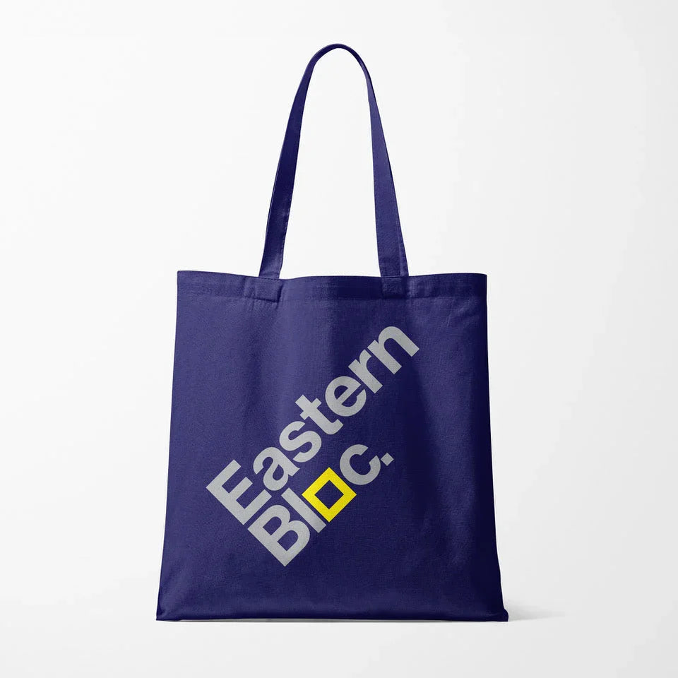Navy blue tote bag with diagonal white text 'Eastern Bloc.' and a yellow square detail on white background