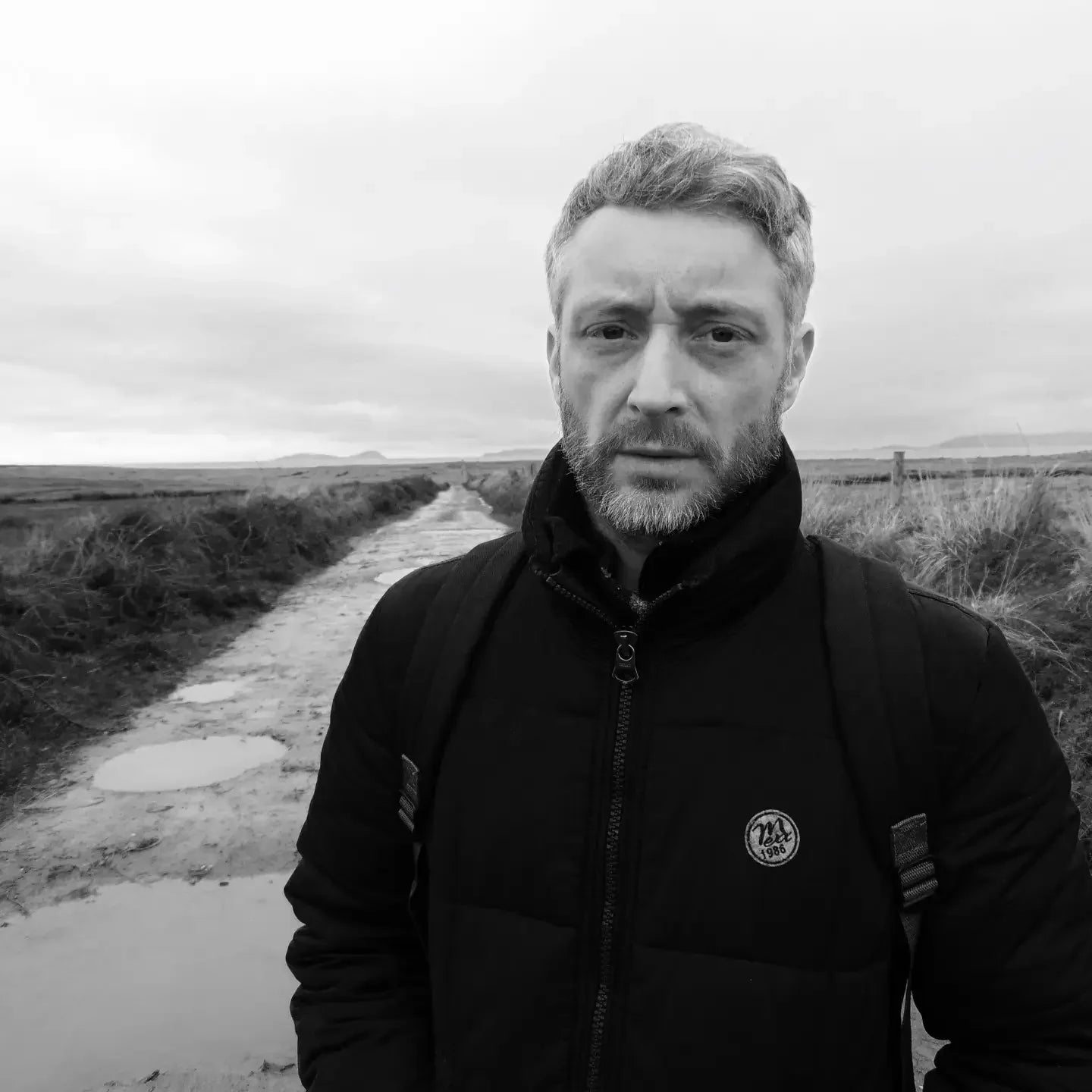 Man in black jacket on rural muddy path, overcast sky, grayscale portrait