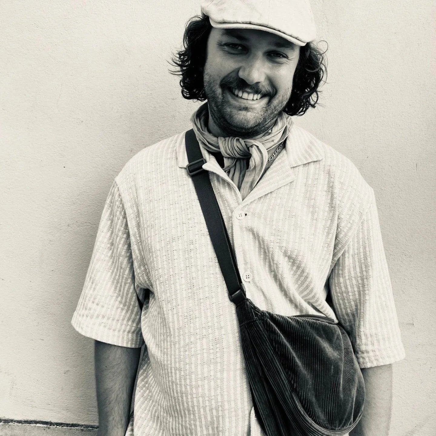 Smiling man in casual shirt, cap, scarf, and crossbody bag against a plain wall