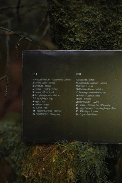 Close-up of a CD tracklist on a black paper in a forest setting with moss and branches.