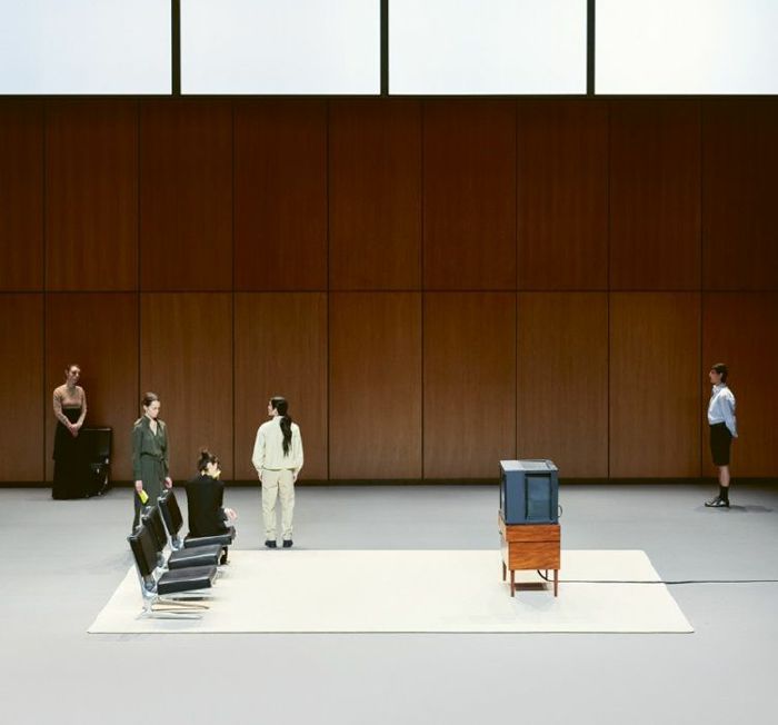 Sculpture of a woman sitting on a chair with a television set for a head, in a minimalistic setting.