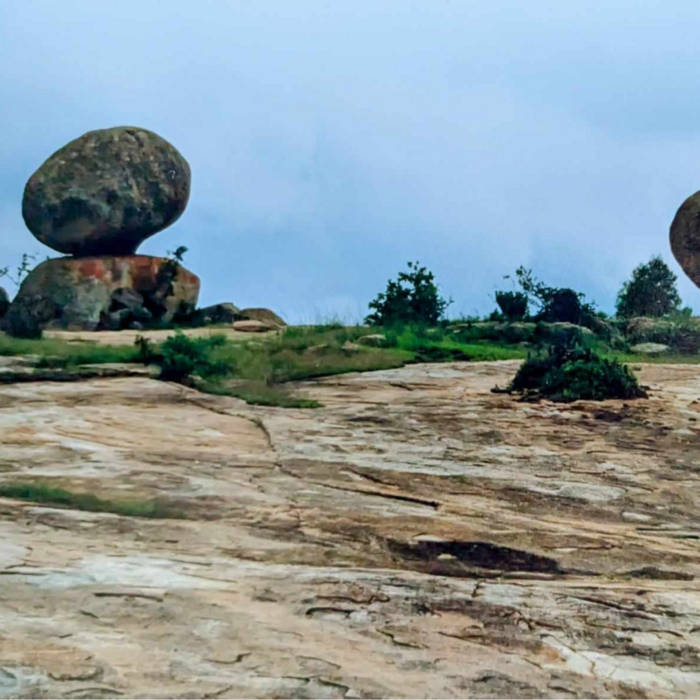 Large round boulder balanced on a flat rock surface with sparse vegetation under a cloudy blue sky